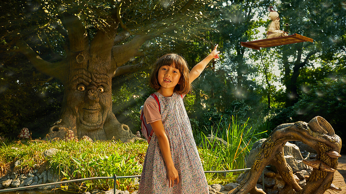 Ein Mädchen zeigt auf den beginnenden Märchenwald im Hintergrund. Ein Baum mit Gesicht beobachtet sie freundlich.
