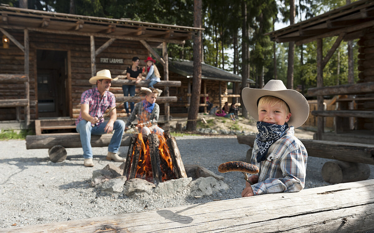Ein Junge mit Cowboy-Hut zeigt eine Grillwurst am Speiß. Im Hintergrund sitz ein Cowboy.