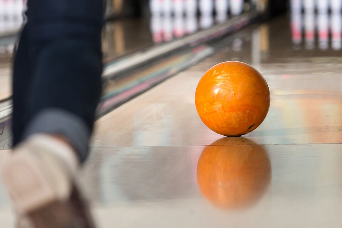 Eine Bowlingkugel rollt auf der Bahn. Der Fuß des Spielers ist unscharf im Vordergrund sichtbar.
