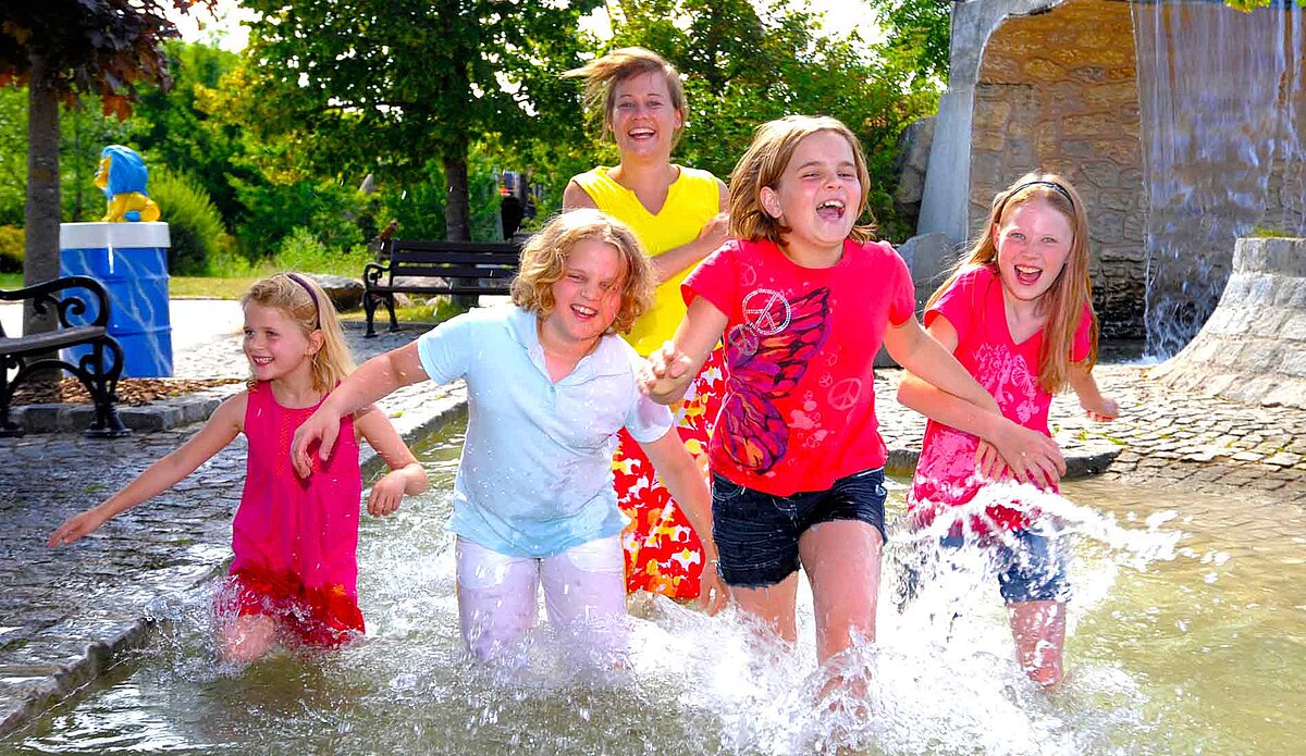 Eine Gruppe Kinder rennt händehaltend durch ein Becken auf dem Wasserspielplatz