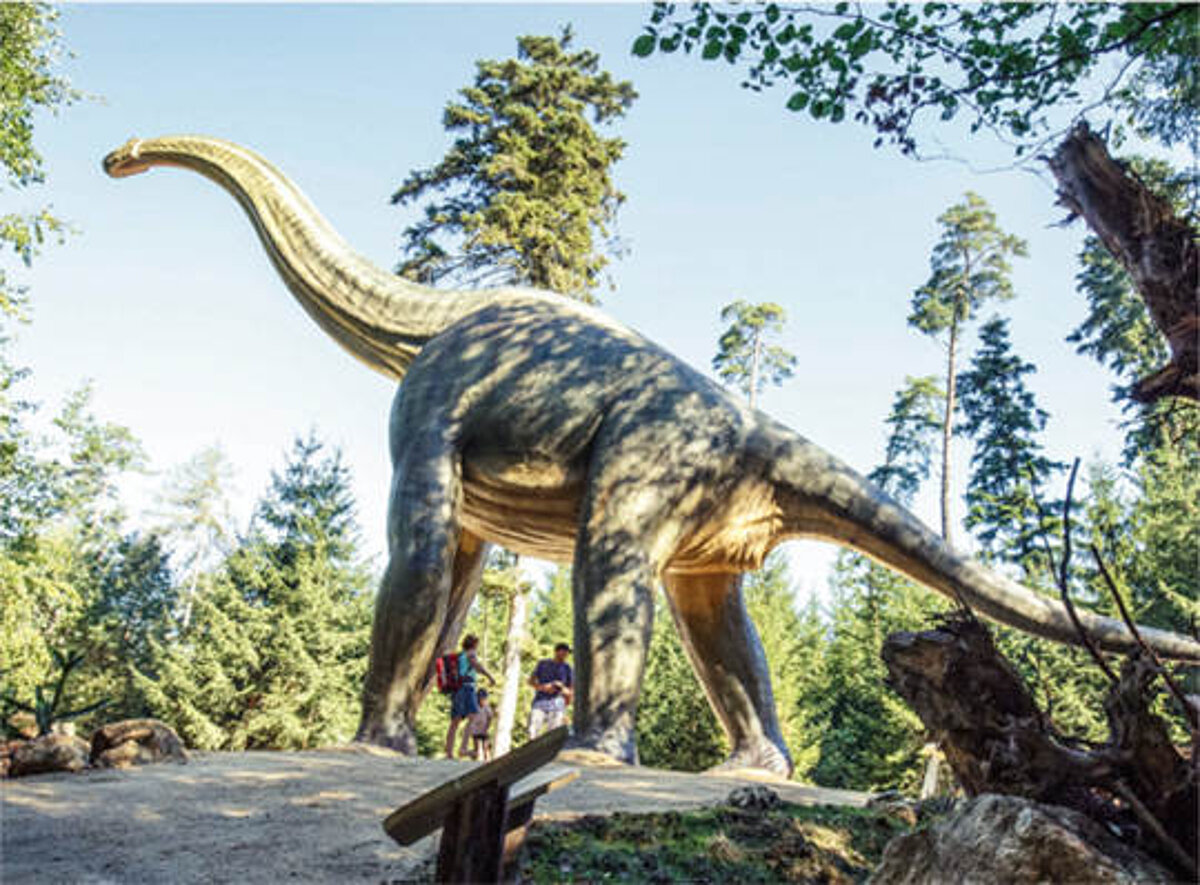 Ein gezichneter Brachiosaurus mit einer Besuchergruppe zu seinen Füßen