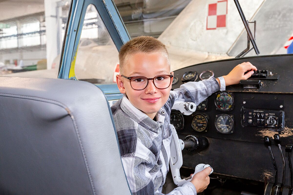 Ein fröhlicher Junge sitzt im Cockpit eines begehbaren Flugzeugs im Hangar H4