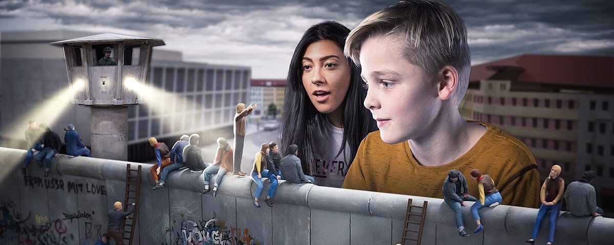 Kinder blciken auf eine Miniatur-Version der Berliner Mauer mit Grenzposten